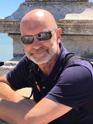 A bald man with a gray beard wearing sunglasses and a navy polo shirt sits outdoors by a stone wall, smiling on a sunny day.