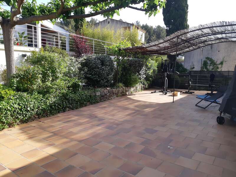 A spacious tiled patio with lush greenery, partial shade from a pergola, outdoor exercise equipment, lounge chairs, and a modern house in the background. Peaceful atmosphere.