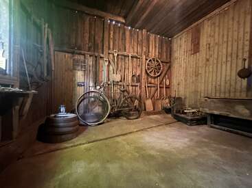 This rustic wooden shed contains vintage tools, an old bicycle, a wagon wheel, barrels, shovels, and other equipment hanging neatly against the walls, illuminated by soft light.