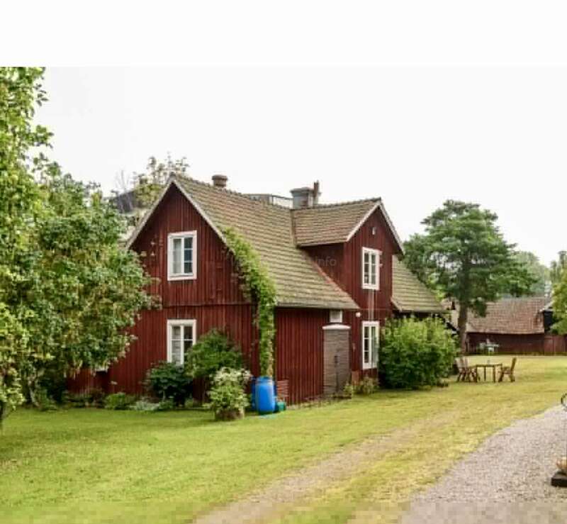 A charming red wooden house with a tiled roof, surrounded by greenery, bushes, and trees. There’s outdoor seating and a gravel path leading alongside it.