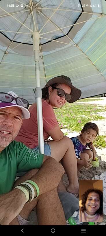 A family relaxes under a large beach umbrella, enjoying the sand. One person joins virtually via video call, smiling warmly in the corner of the photo.