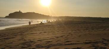 A serene beach at sunset, golden sunlight reflecting off the water. Silhouetted people stroll along the shore, distant waves, and footprints scattered across the sand.