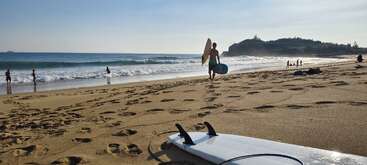 A sunny beach scene with surfers and people enjoying the waves. A surfboard lies on the sand, footprints everywhere, and cliffs rise in the distance.