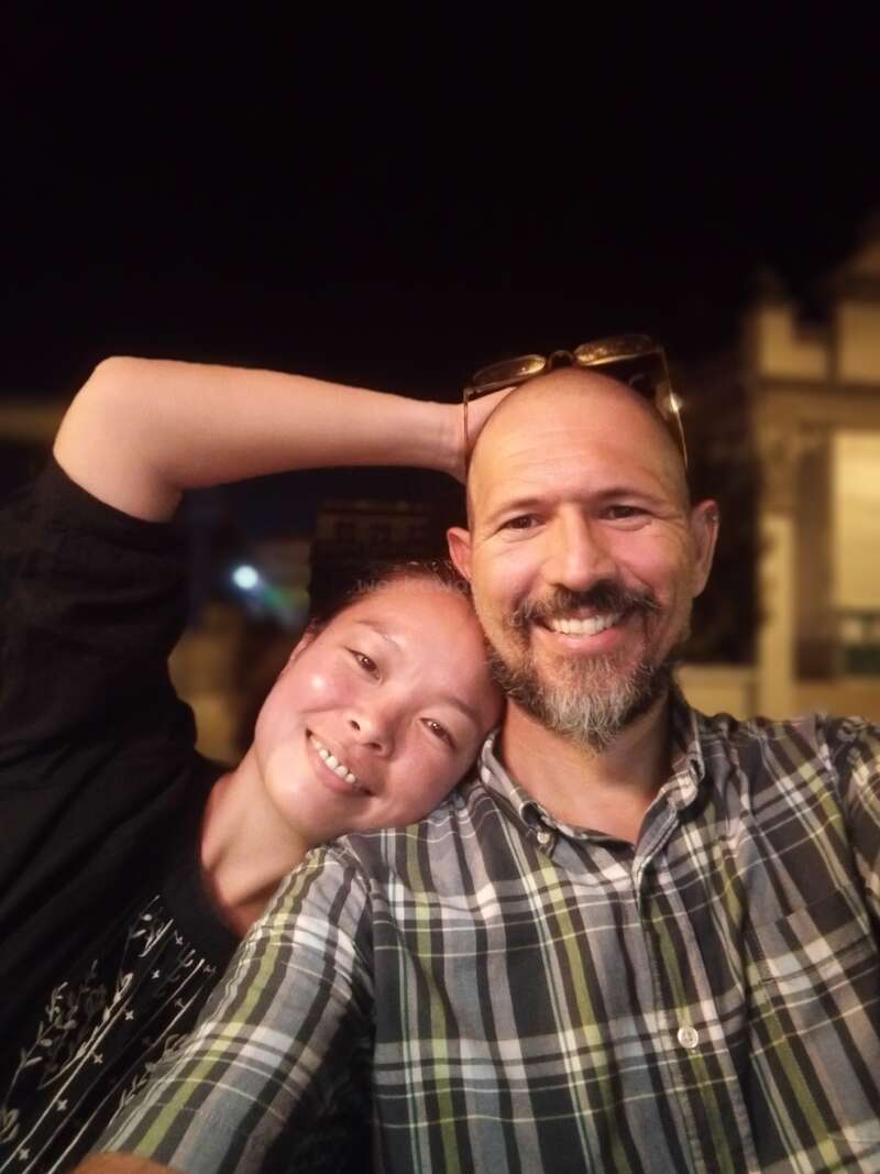 The image depicts a man and woman posing for a photo, with the woman leaning on the man\'s shoulder and her arm raised behind his head.