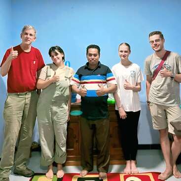 Five people stand indoors against a light blue wall, smiling and giving thumbs up. They are barefoot, dressed casually, and appear to be posing for a group photo.