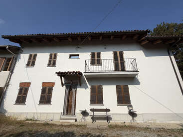 Uma casa de dois andares com paredes brancas, persianas marrons, telhado de madeira, varanda pequena e alpendre na entrada. Área externa com um banco e duas cadeiras na frente.