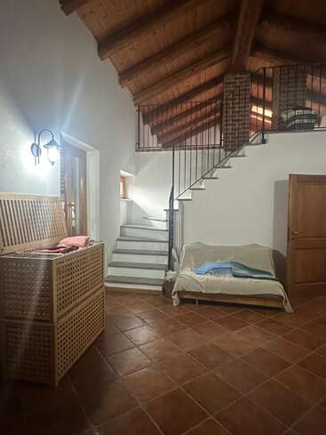 This cozy room features terracotta tile floors, wooden ceiling beams, a staircase leading to a loft, covered sofa, wicker storage, and warm, ambient lighting.