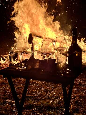 Uma fogueira acesa ilumina uma mesa rústica de madeira com taças de vinho, uma garrafa e livros, criando um clima aconchegante e convidativo para uma reunião noturna ao ar livre.