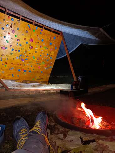 Uma pessoa relaxa à noite perto de uma pequena fogueira, usando sapatos de escalada, com uma parede de escalada ao ar livre bem iluminada e um grande dossel acima dela ao fundo.