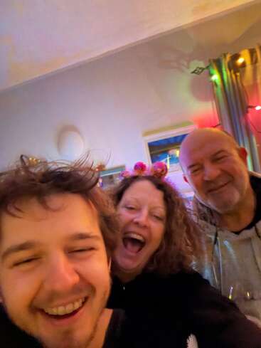 Três pessoas sorriem alegremente para uma selfie dentro de casa. O clima é festivo, com luzes coloridas e expressões animadas, sugerindo uma reunião ou comemoração divertida e feliz.