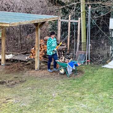 A person stands outdoors near stacked firewood, using an axe to cut logs. A wheelbarrow with clothes is nearby, under a wooden shelter, surrounded by trees.