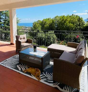 Un acogedor patio exterior cuenta con muebles de mimbre, una mesa de cristal, una alfombra estampada y un gato descansando. Los frondosos árboles y las vistas al mar completan la serena escena.