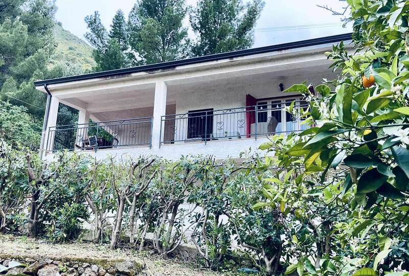 Una casa blanca con un amplio balcón cubierto, rodeada de exuberante vegetación y arbustos recortados. Sillas rústicas, fondo montañoso, árboles altos y un apacible ambiente natural.