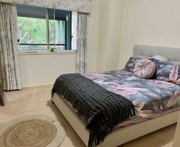 Une chambre à coucher confortable avec un lit double doté d'une literie tropicale, d'oreillers décoratifs, d'un jeté texturé, de rideaux floraux, d'une vue sur la fenêtre et d'un tapis rond en toile de jute.