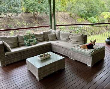 Un patio extérieur confortable avec un canapé sectionnel en osier, un coussin vert, une table en verre et un petit chien portant un pull qui se prélasse sur le coussin.