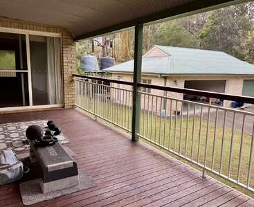 Une terrasse en bois couverte donne sur une cour herbeuse et un garage. Du matériel d'exercice et un sac de sport sont posés sur un tapis près des portes coulissantes en verre ; des arbres l'entourent.