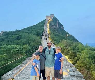 Une famille de quatre personnes pose joyeusement sur la Grande Muraille de Chine au crépuscule, entourée de verdure luxuriante et de chemins illuminés, avec des sourires et des vêtements décontractés.