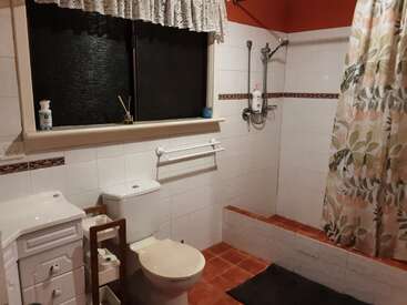 Esse banheiro tem vaso sanitário, pia, janela com cortina, chuveiro acessível com barras de apoio, cortina de chuveiro floral, piso de azulejo e paredes com azulejos brancos e detalhes em vermelho.
