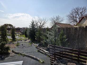 A sunny backyard garden features young trees, garden beds covered with black plastic, a wooden fence, rustic log borders, and scattered gardening supplies. Houses and bare trees behind.