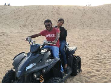 Das Bild zeigt einen Mann und eine Frau, die mit einem ATV in der Wüste fahren, mit einer Sanddüne hinter ihnen und Menschen, die in der Ferne auf der Düne stehen.