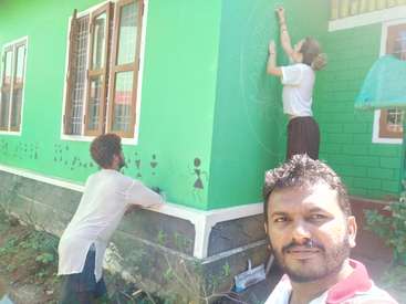 Das Bild zeigt zwei Personen, die ein grünes Haus mit braunen Motiven streichen, während ein Mann im Vordergrund ein Selfie macht. Die Frau steht auf einer Leiter.