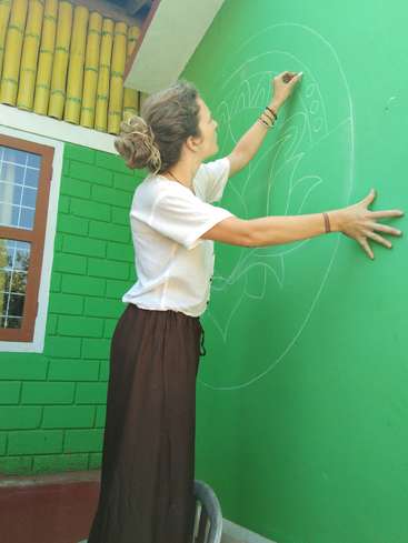 Eine Frau mit einem Dutt malt mit Kreide auf eine grüne Wand. Sie trägt ein weißes Hemd und eine braune Hose.