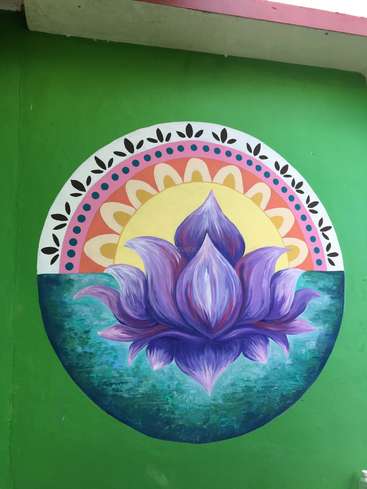 Das Bild zeigt ein lebendiges Wandgemälde mit einer lila Lotusblume auf einer grünen Wand vor einem Sonnen- und Regenbogenmotiv mit einem weißen Rand aus Blättern.