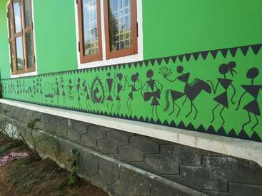 Das Bild zeigt eine leuchtend grüne Wand mit schwarzer Warli-Stammeskunst, auf der Menschen, Tiere und geometrische Muster abgebildet sind, vor einem weiß gerahmten Fenster und einem steinernen Fundament.