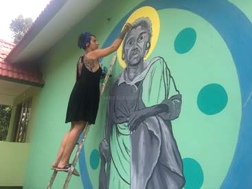 Eine Frau mit Tattoos und blauen Haaren steht auf einer Leiter und malt ein Wandgemälde von einer Person mit einem gelben Heiligenschein und blauen Punkten auf eine grüne Wand.