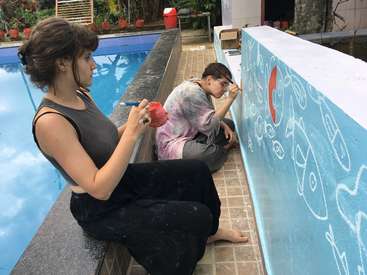 Zwei Frauen malen mit Pinseln und Farbdosen ein Wandbild an eine Wand neben einem Pool. Die Wand ist blau mit weißen Mustern.