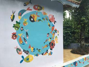 Das Bild zeigt ein lebhaftes Wandgemälde mit Cartoon-Fischen auf einer weißen Wand, vor einem Hintergrund aus üppigem Grün und einem Baum.