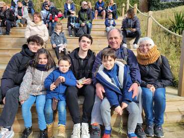 A multigenerational family sits together on wooden steps outdoors, smiling and enjoying a sunny day. Other families sit in the background, adding to the lively atmosphere.
