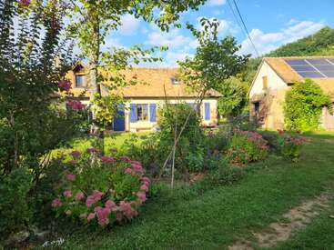 A charming countryside house with blue shutters sits amidst vibrant flower gardens. Nearby, an outbuilding with solar panels reflects sustainability. Lush greenery and sunshine complete the scene.