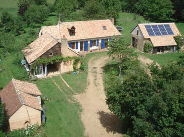 Aerial view of a rustic countryside property with three tiled-roof buildings, solar panels, lush greenery, dirt pathways, and surrounding trees under a bright sky. Peaceful scene.