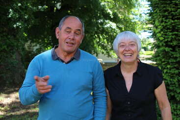 A man in a blue sweater and a woman in a black shirt are smiling outdoors, surrounded by greenery and trees on a sunny day.