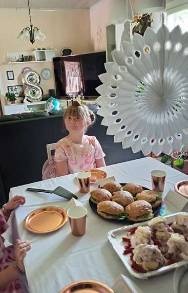 Uma menina usando uma coroa senta-se em uma mesa decorada com sanduíches, sobremesas, pratos dourados e xícaras, comemorando o que parece ser um oitavo aniversário.