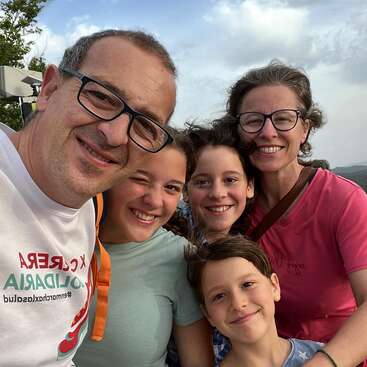 A happy family of five, smiling outdoors on a partly cloudy day. Everyone looks cheerful and close, enjoying a moment together for a group selfie.