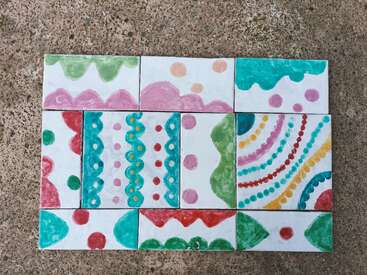 Nove azulejos pintados à mão dispostos sobre uma superfície de concreto, com padrões coloridos com pontos, linhas onduladas e conchas em verde, rosa, turquesa, vermelho e amarelo.
