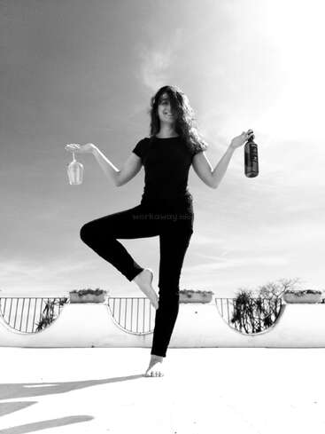 The image depicts a woman in a yoga pose, holding a wine glass and bottle, with a serene background featuring a balcony and a tree.