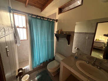 Este cuarto de baño cuenta con una ducha con una cortina azul, un inodoro, un lavabo con espejo, un techo de madera, una estantería con artículos de aseo, una ventana y da a un dormitorio.