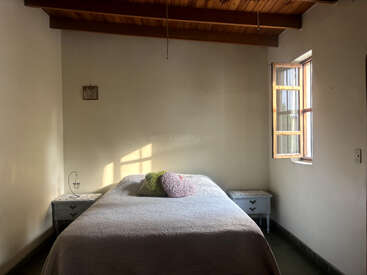 Un dormitorio sencillo con techo de madera, cama de matrimonio, almohadas, dos mesitas de noche, una lámpara pequeña y una ventana abierta por la que entra la luz del sol. Ambiente acogedor.