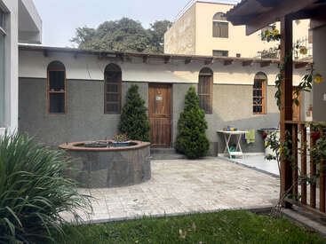 Esta imagen muestra un pequeño patio con un pozo de piedra, dos arbustos de hoja perenne, una puerta de madera, ventanas en arco, suelo de baldosas y ropa tendida al aire libre.