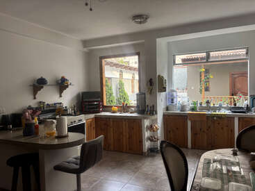 Se trata de una cocina acogedora y luminosa, con armarios de madera, ventanas grandes, diversos electrodomésticos, una barra de desayuno, plantas de interior y una mesa de comedor que se vislumbra parcialmente al fondo.