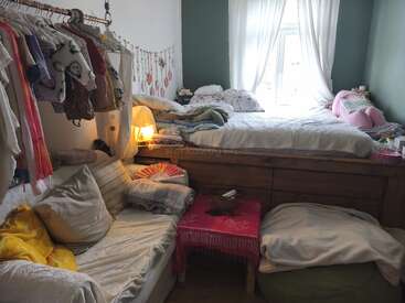 Se trata de un dormitorio acogedor y bien iluminado, con una cama elevada junto a la ventana, ropa de cama mullida, un sofá, ropa colgada, cojines y cálidos detalles decorativos, todo ello bañado por una luz tenue.
