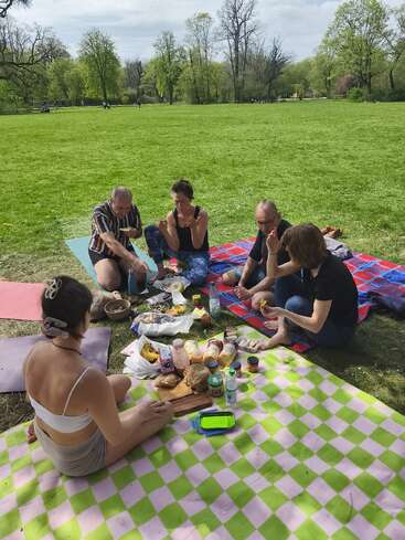 Cinco personas están sentadas sobre mantas de picnic en un parque soleado, rodeadas de comida y bebida, disfrutando de una comida juntos bajo el cielo despejado y los árboles frondosos.