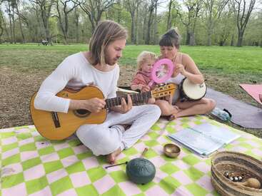 Un hombre toca la guitarra mientras una mujer con un niño toca una pandereta rosa y un tambor sobre una manta de picnic en un tranquilo y frondoso parque.