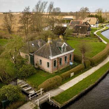 Une charmante maison en briques se dresse au bord d'un canal, entourée de haies verdoyantes, d'arbres et d'un bateau. D'autres maisons au toit de chaume bordent le cours d'eau paisible tout près.
