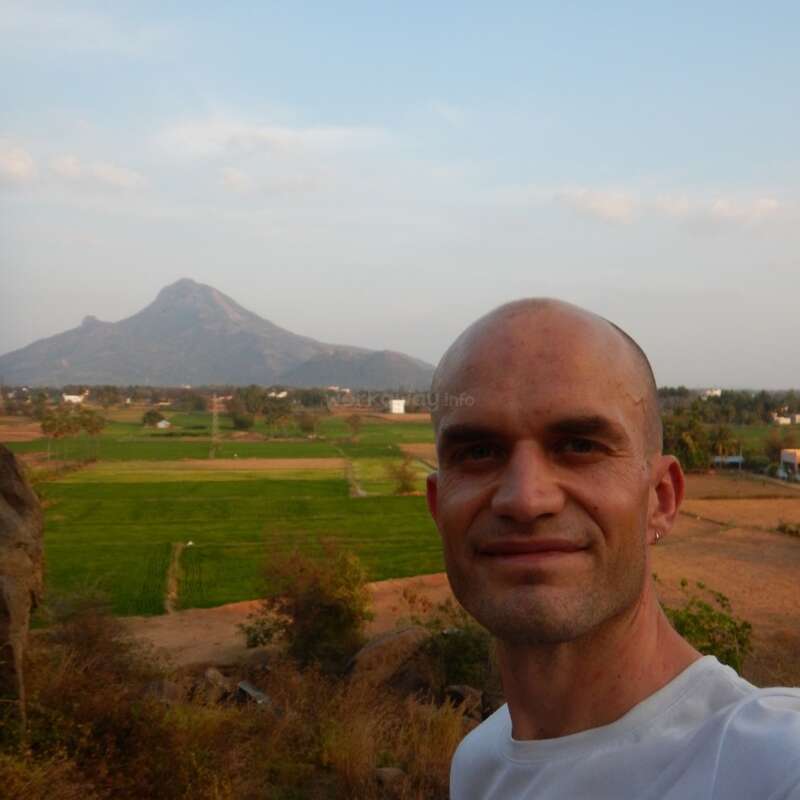 Ein lächelnder Mann macht ein Selfie vor einer malerischen Landschaft mit weiten Feldern, vereinzelten Häusern, Bäumen und einem Berg in der Ferne unter strahlend blauem Himmel.