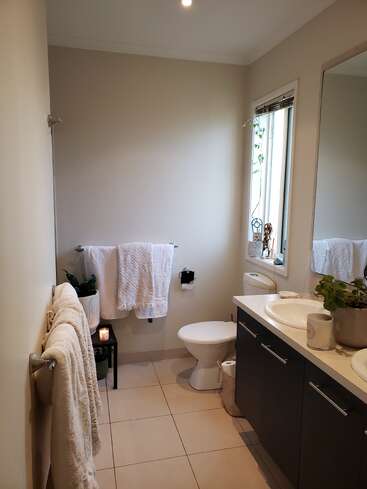 This image shows a modern, tidy bathroom with light tiles, white towels, a toilet, plants, a large mirror, countertop sinks, a window, and soft lighting.