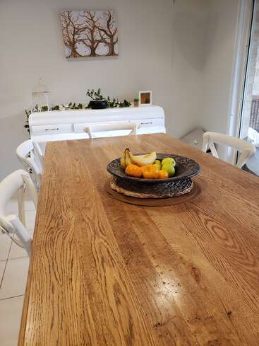 A wooden dining table with white chairs, centerpiece fruit bowl, and natural light. In the background, a white sideboard, plants, and tree artwork decorate the space.